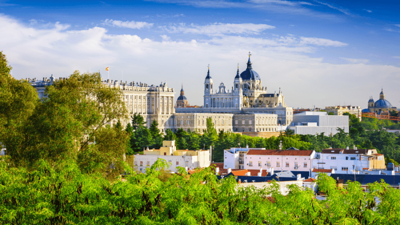 Madrid skyline featuring the Royal Palace and Almudena Cathedral, viewed from a green park under a clear blue sky.