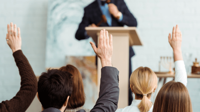 A view from behind of people raising their hands to bid at a property auction, with the auctioneer at a podium in the background.