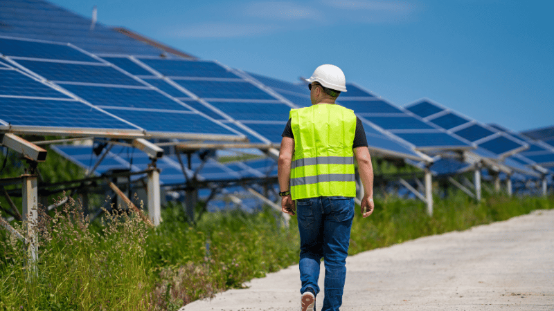 An energy management professional in a hard hat and safety vest inspects a solar farm, representing a business's investment in sustainable energy.