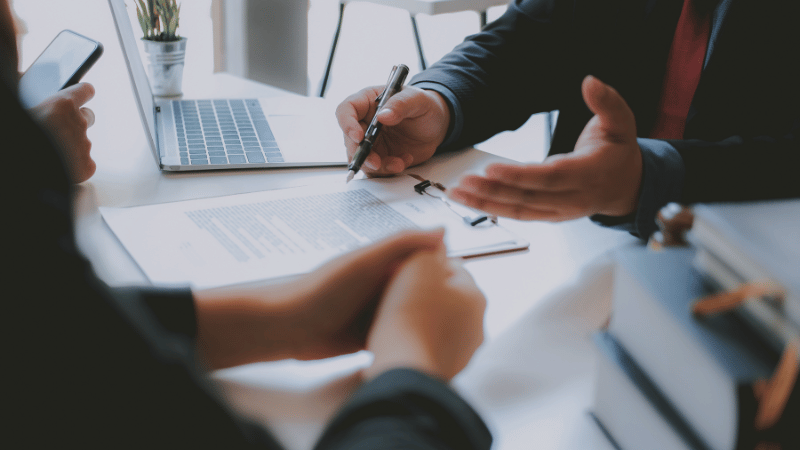 A business person in a suit sits at a desk, pointing to a document on a clipboard while another person's hands are clasped, representing a financial consultation.