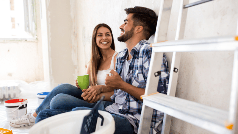 A happy couple taking a break with coffee during their house renovation project, sitting on the floor surrounded by paint pots and a ladder.