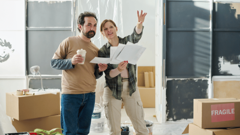 A smiling couple in a room under renovation, reviewing architectural plans and discussing their home improvement project.