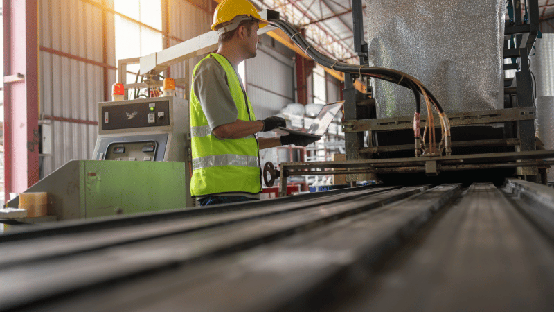 A manufacturing worker in a hard hat and safety vest uses a laptop to operate a production line, representing the use of asset finance to upgrade machinery