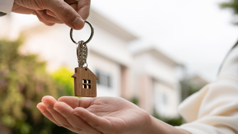 A person handing house keys with a wooden house-shaped keyring to a new homeowner, symbolizing the successful outcome of finding one of the mortgage best buys.