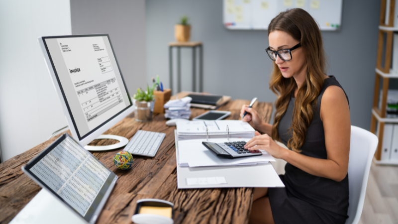 A woman wearing glasses in an office, using a calculator and reviewing financial documents with an invoice visible on a computer screen, representing self-assessment tax calculations.