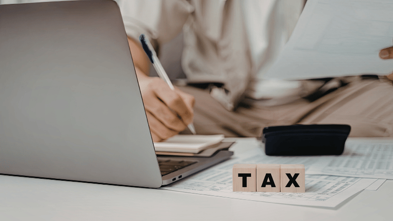A person working on a laptop to calculate their tax bill, with wooden blocks spelling out the word TAX on the desk in front.