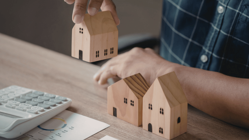A person arranging small wooden model houses on a desk with a calculator and financial chart, symbolizing planning and forecasting for the UK property market.
